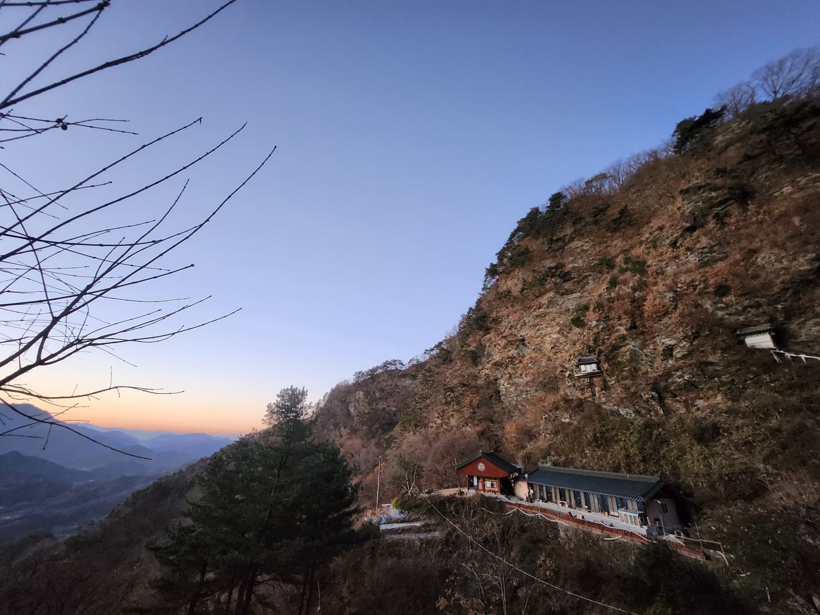 Before sunrise: the temple's red main hall and long residence at the foot of a dark cliff, with two small shrines visible on the rock face