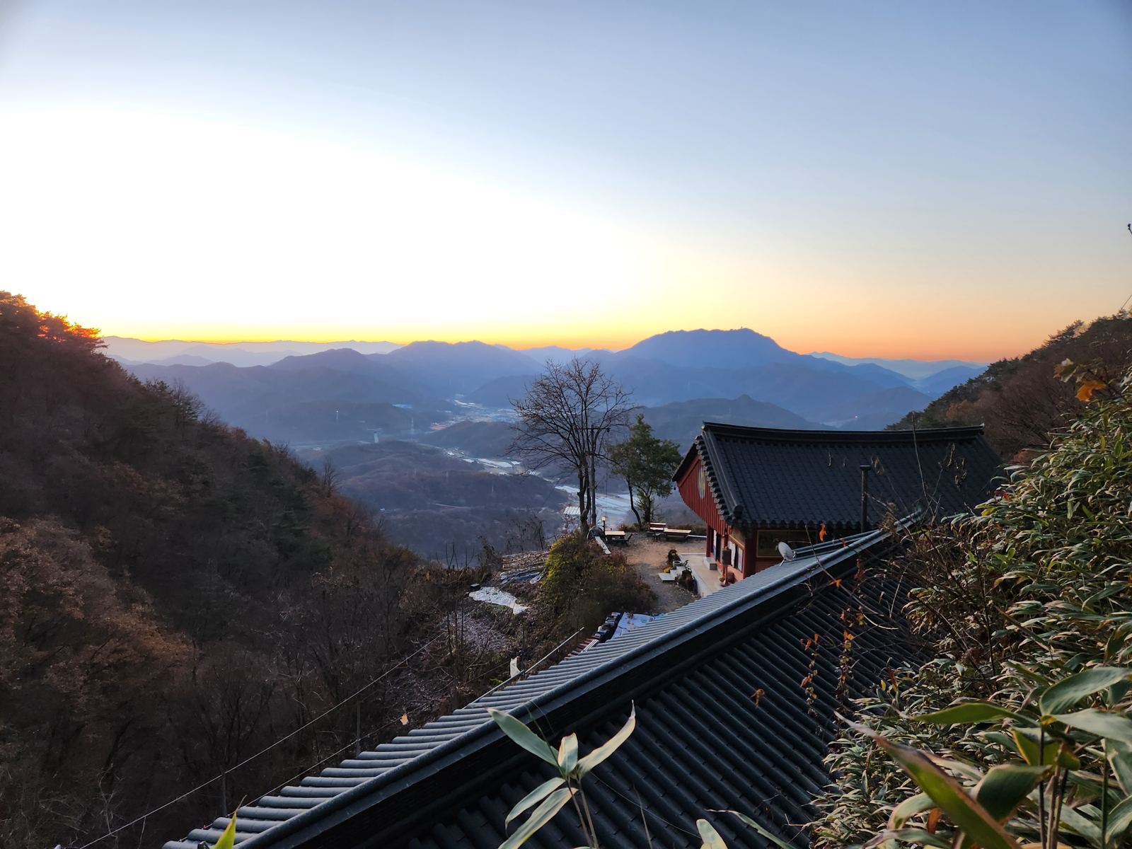 The temple's tiled roofs and red main hall at dawn, with the village at the foot of the mountain and a distant flat-topped peak against an orange horizon