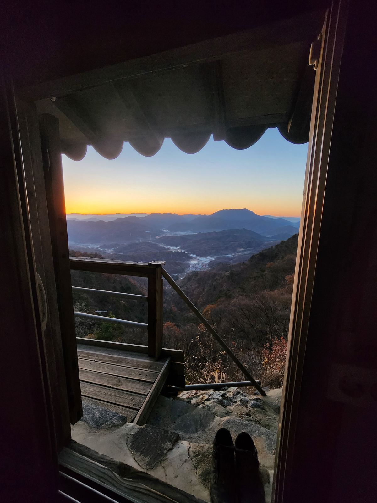 View from inside the shrine doorway: a wooden balcony and a valley of layered mountains under an orange dawn sky, a pair of shoes on the stone step