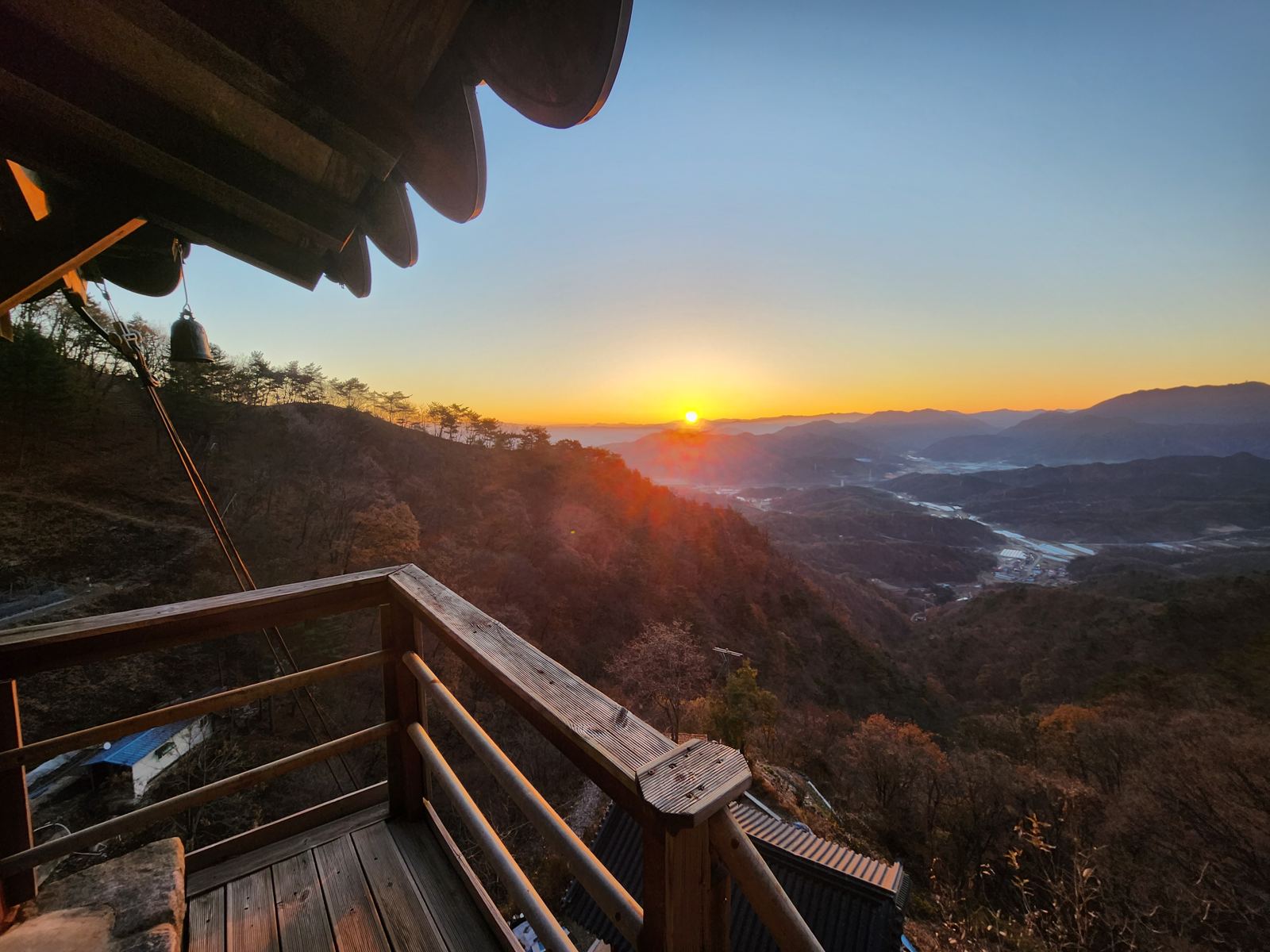 Sunrise seen from the shrine's wooden balcony, a bell hanging from the eaves and a valley of mountains below