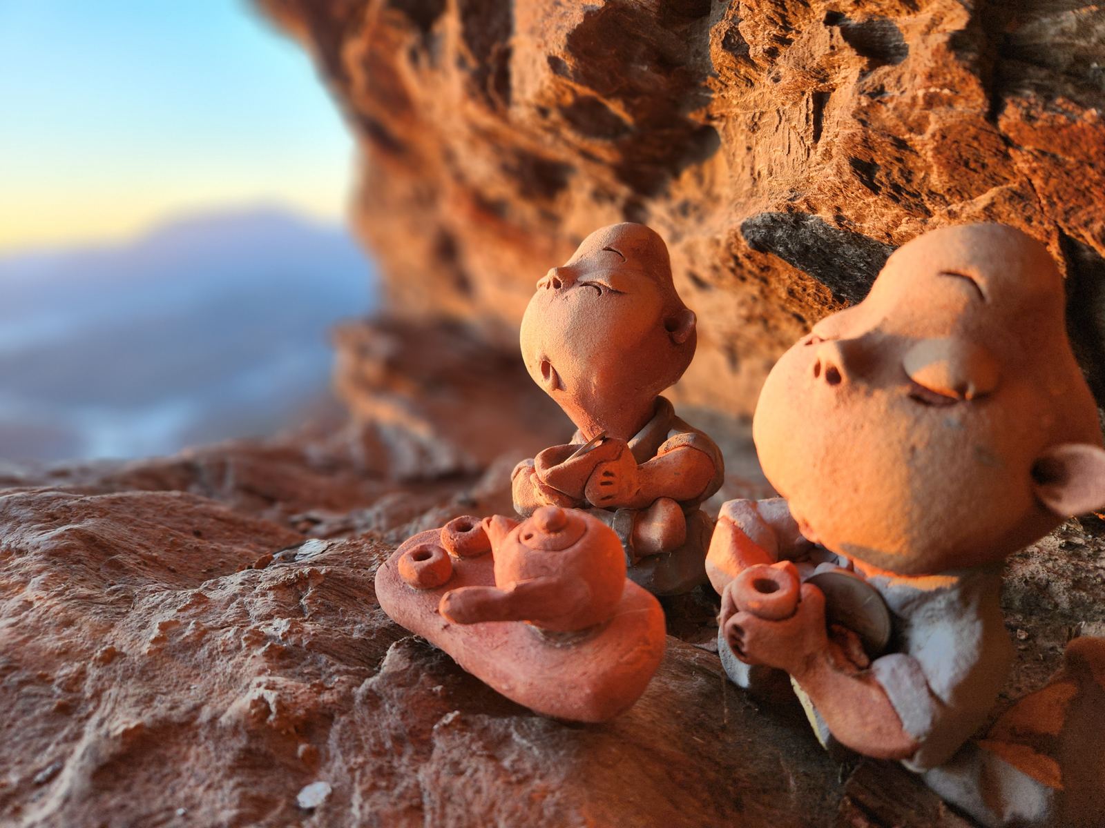 Two clay figurines of child monks with a teapot and cups on a rock ledge, faces turned up, lit by the morning sun