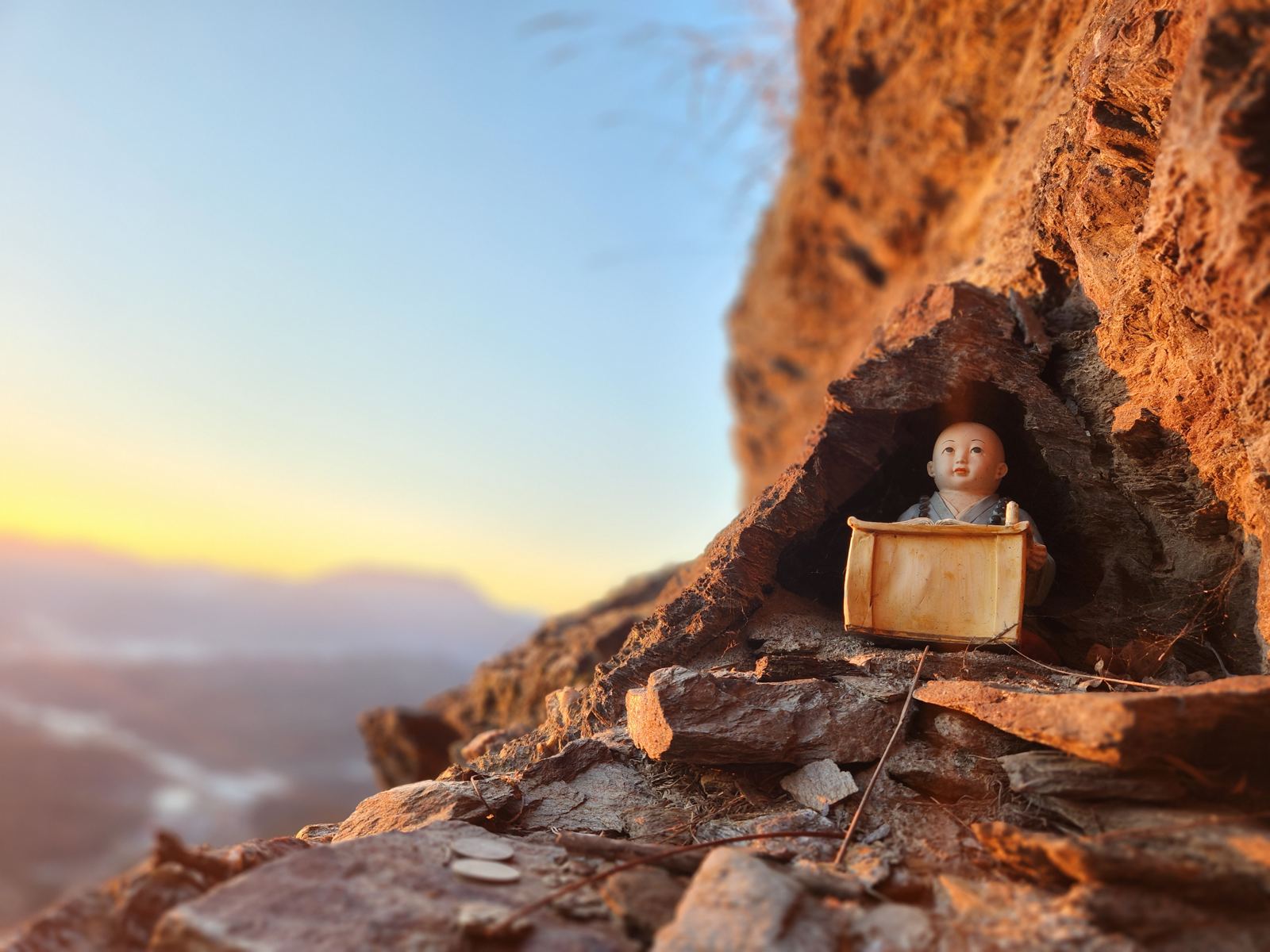 A small figurine of a child monk reading a scroll inside a hollow in the orange rock, coins on the ledge in front
