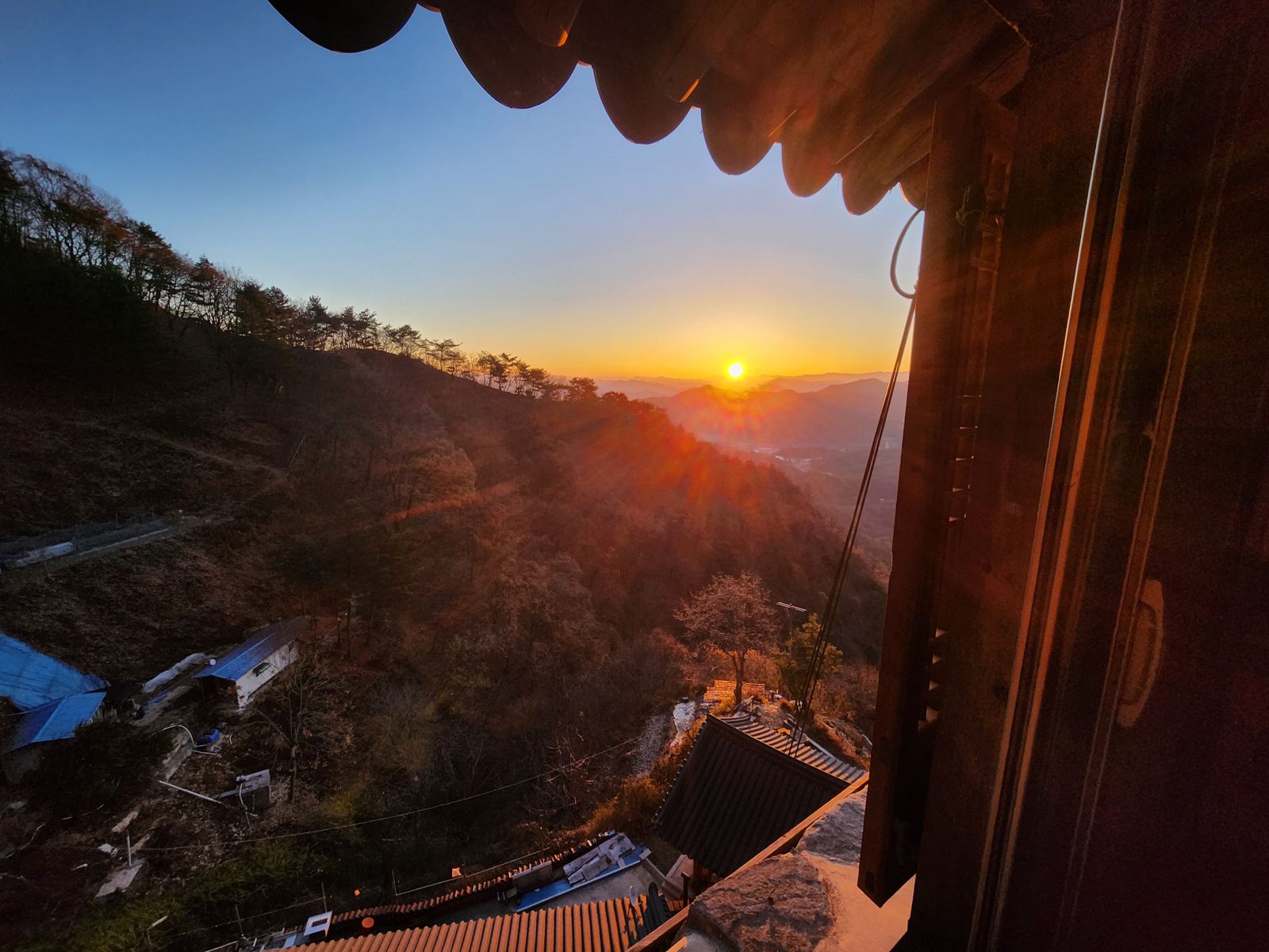 The sun over layered ridges seen past the shrine's eaves and window frame, the temple roofs far below