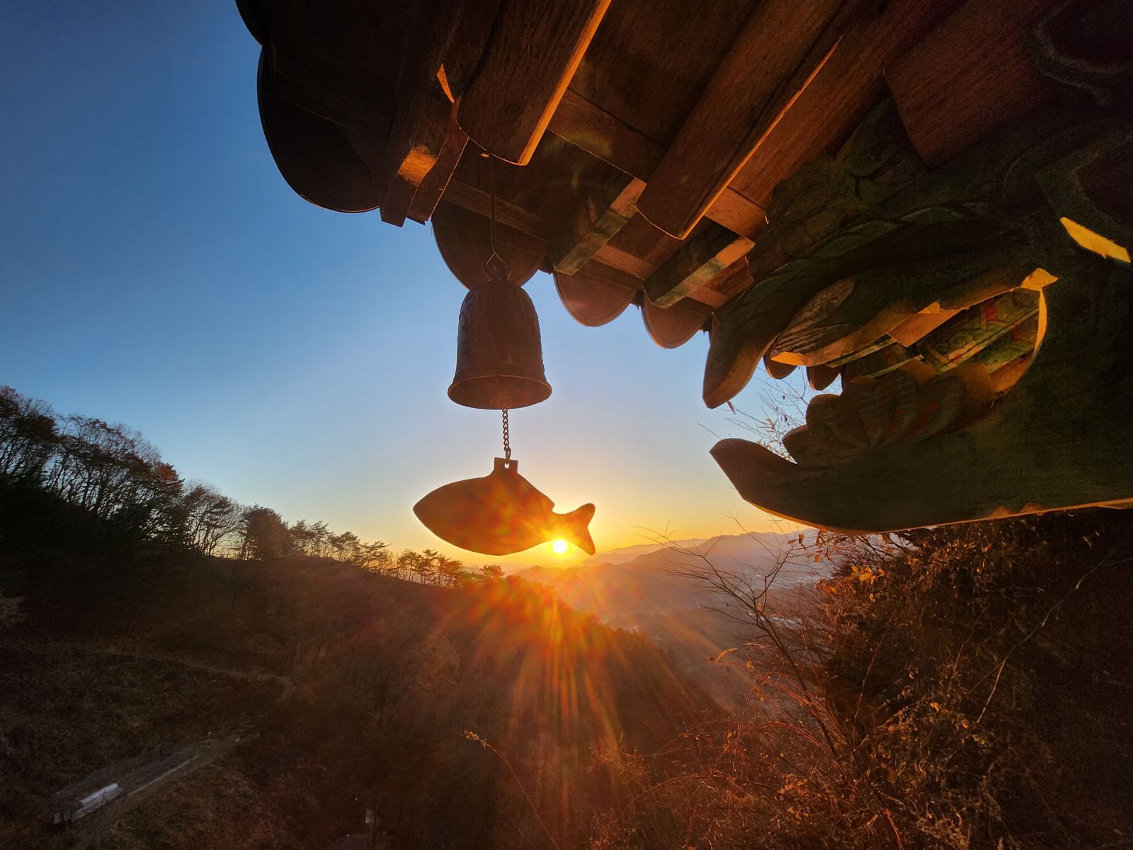 A bronze wind bell with a fish-shaped clapper silhouetted against the rising sun under painted eaves