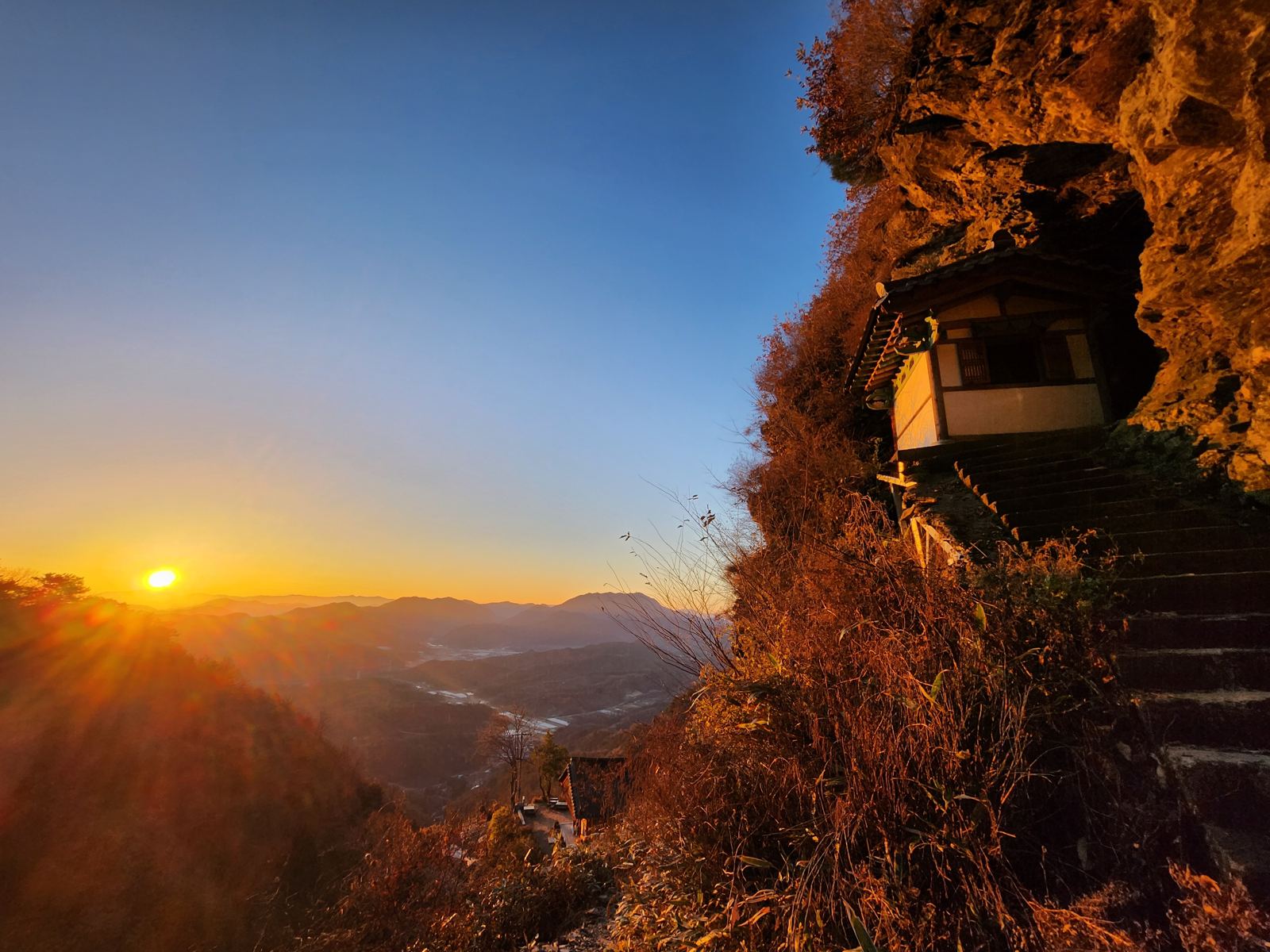 A small shrine built into a hollow of the cliff, with the sun rising over layered mountains to the left