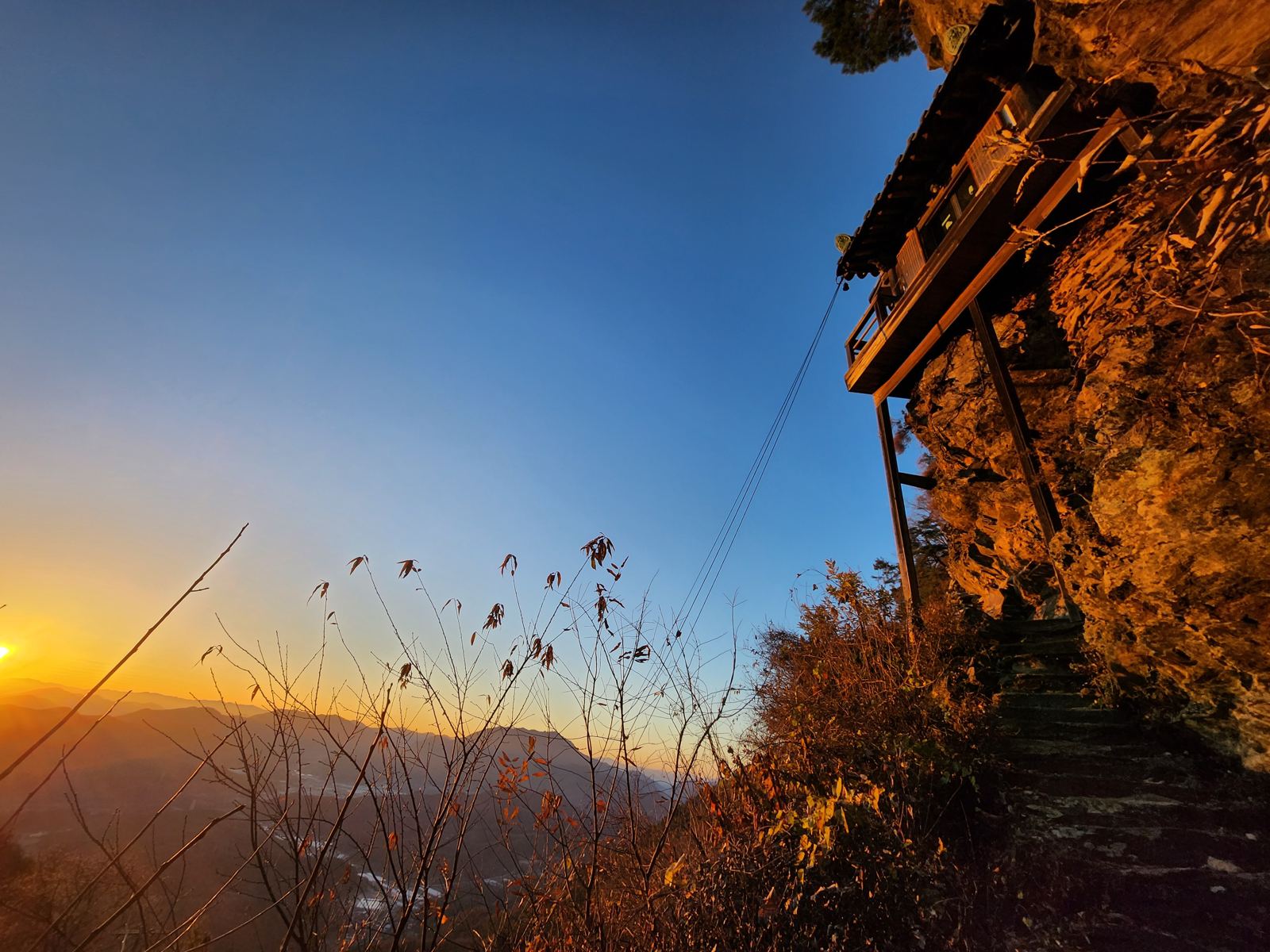 Looking up at a small shrine on wooden stilts against the cliff, a deep blue sky and a valley at sunrise behind