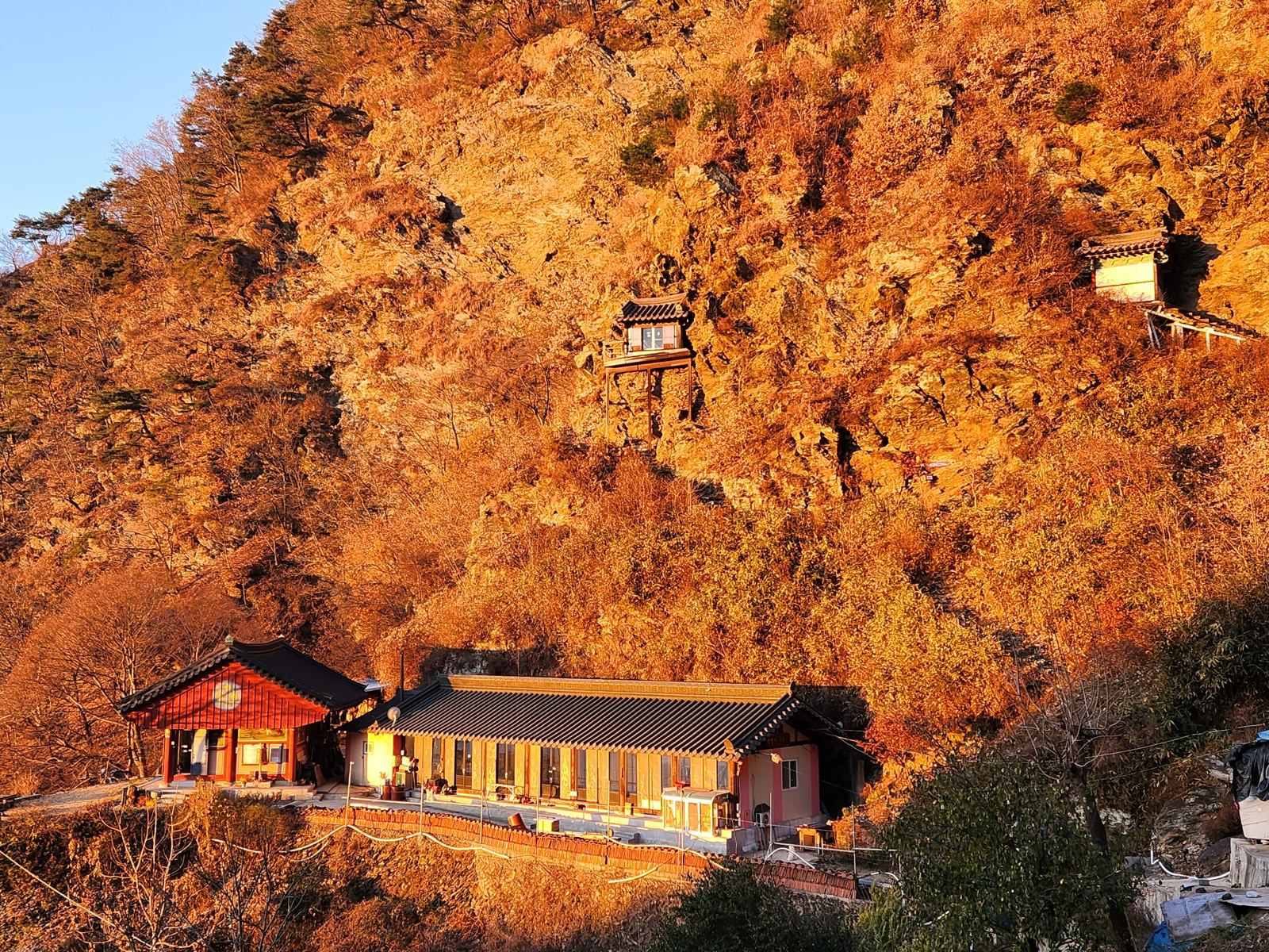 Two tiny shrines fixed to a sunlit orange cliff above the roofs of Gujeolsa temple