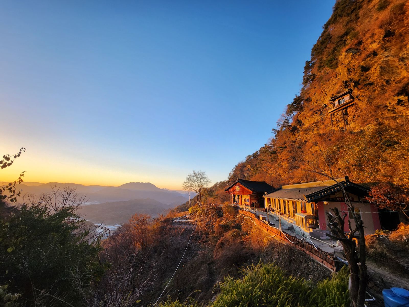 Wide view in full morning light: the temple buildings on a narrow ledge, the stilt shrine on the cliff above, the valley and mountains to the left