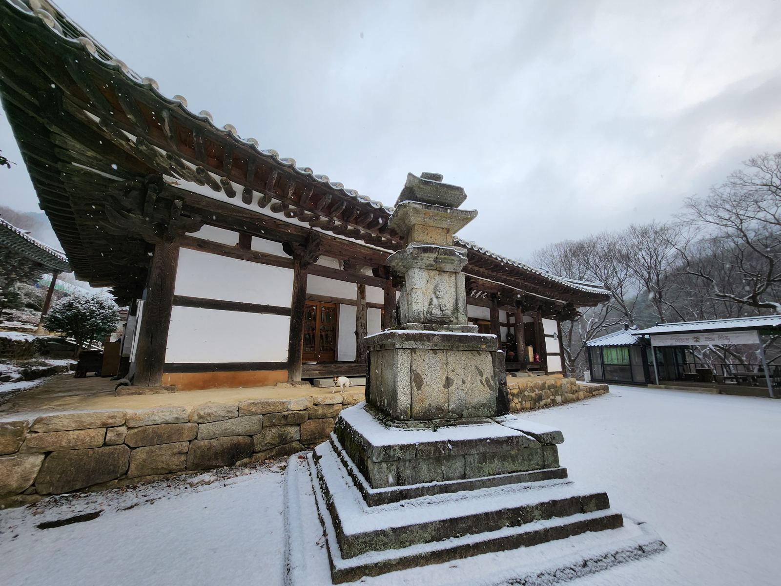 A weathered three-tier stone pagoda dusted with snow in front of a low hermitage building