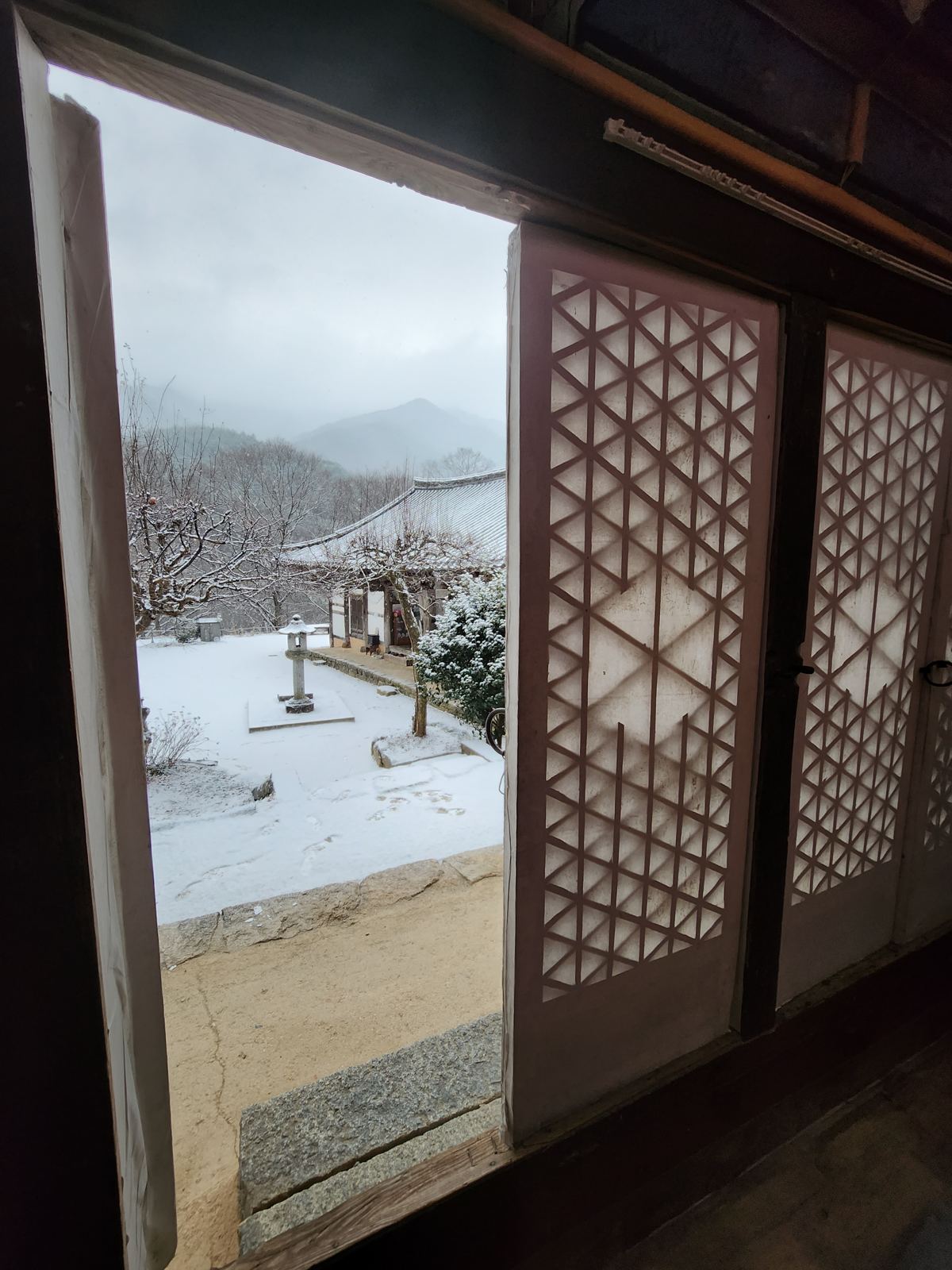 View through an open lattice door onto a snowy courtyard, a stone lantern and misty mountains