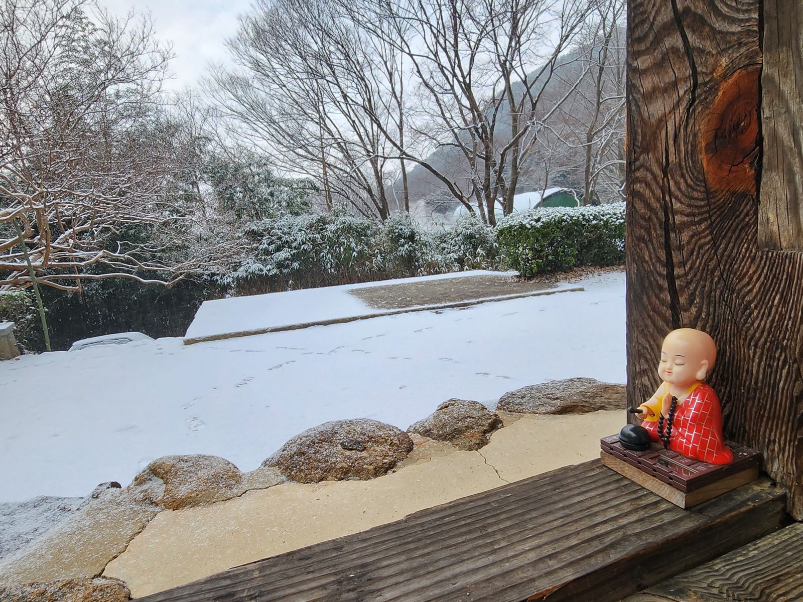 A small figurine of a young monk with a wooden moktak on a ledge, a snowy garden beyond