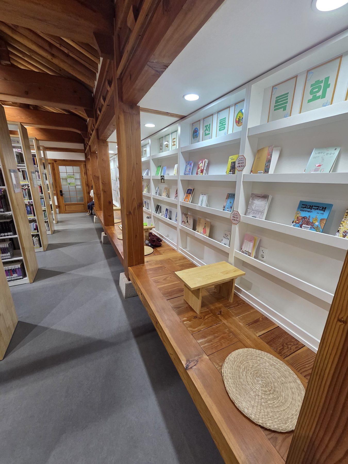An indoor wooden veranda with straw cushions and a low table, beside shelves of books on Korean tradition