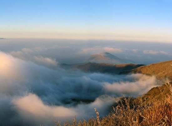 In low golden light, cloud flows down the valleys below the ridge, with one peak rising above it in the distance