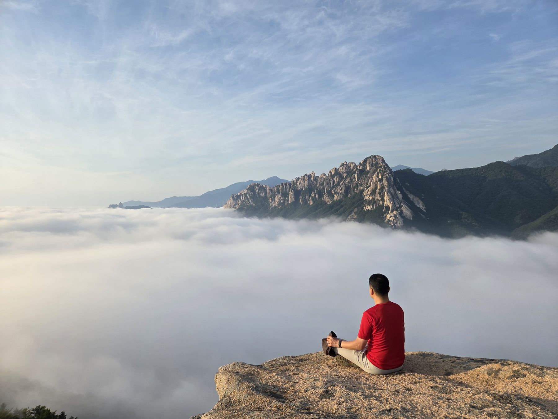 A hiker in a red shirt sits on the edge of the summit rock, facing Ulsanbawi above a sea of clouds