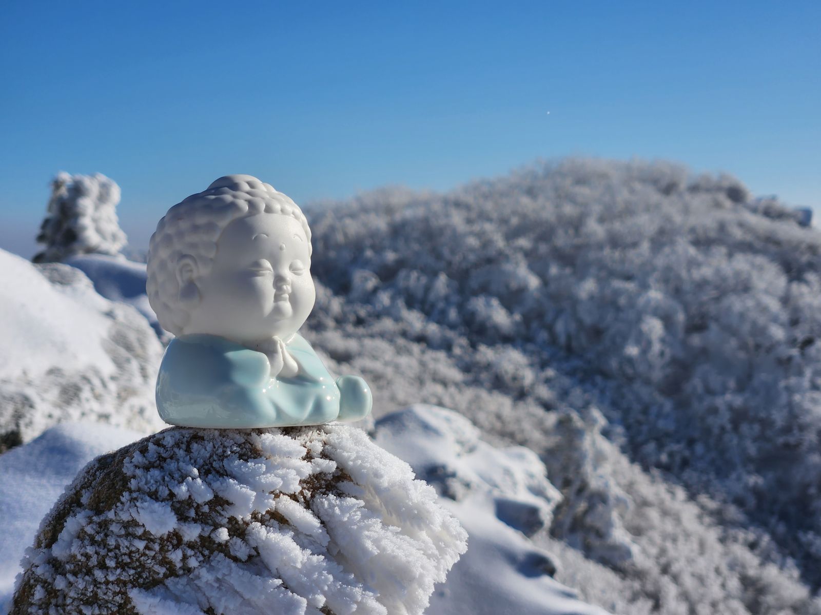 A small ceramic monk figurine set on a frosted rock, with the white frozen ridge behind it
