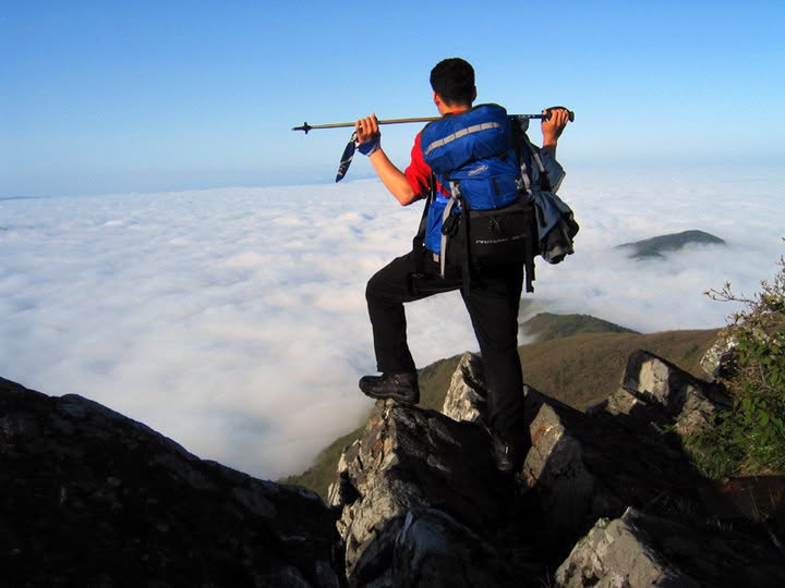 A hiker with trekking poles across his shoulders stands on a rock, looking out over an endless sea of clouds with a peak rising above it