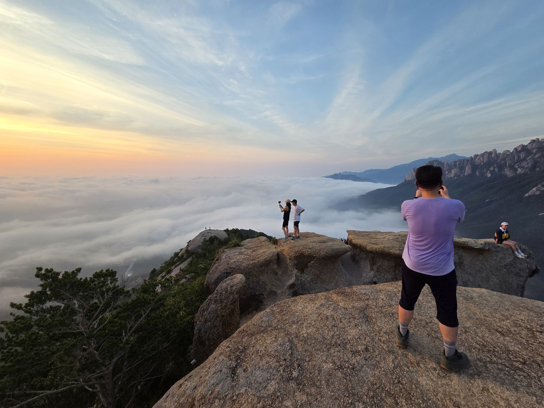 Hikers photograph the sunrise from granite ledges above a sea of clouds, with Ulsanbawi on the right