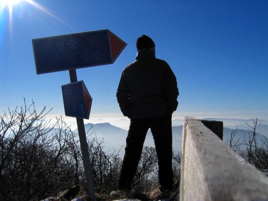 A hiker in silhouette beside the summit signpost, looking out over winter ridges against the sun