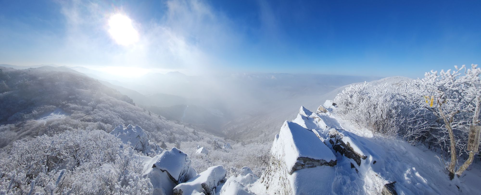 Beside the Duwibong summit post, rime-covered shrubs and snowy rocks open onto winter ridges under a hazy sun